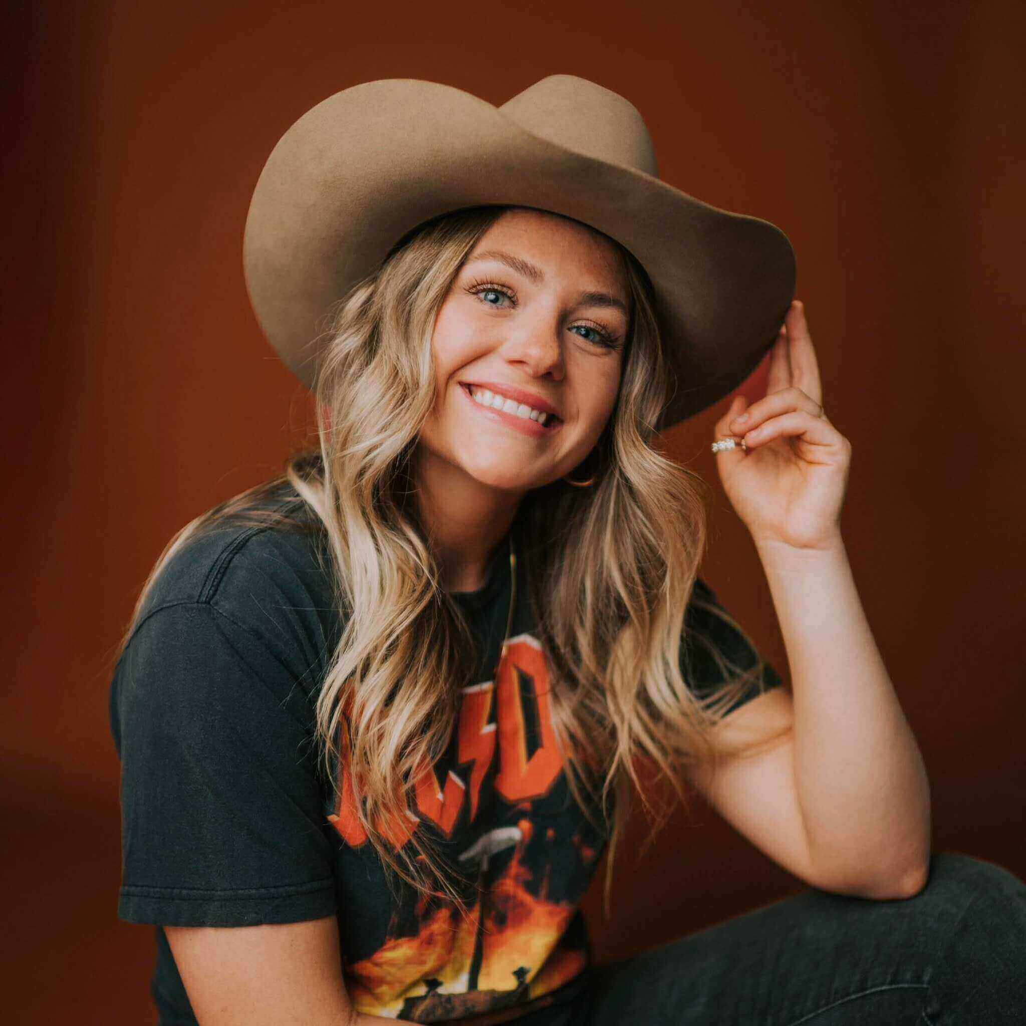 A girl with blonde hair wearing a browen cowboy hat and grey t shirt with a red background