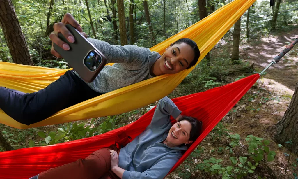 A couple of women laying on hammocks and taking a selfie in the woods.